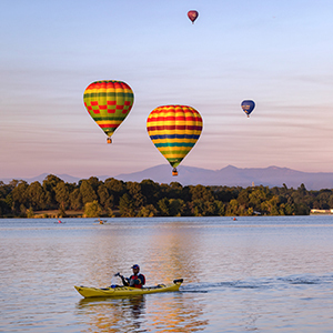 A person kayaking on Lake Burley Griffin at sunrise, as colourful hot air balloons drift across the sky