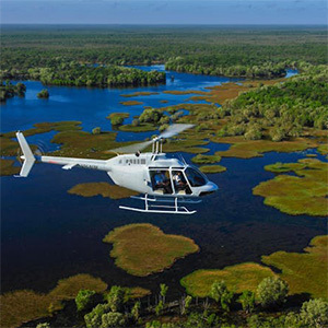 Helicopter soars over wetlands and lush green wilderness landscape.