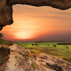 Vast landscape unfolds: orange sunset, green grass, and rocky viewpoint.