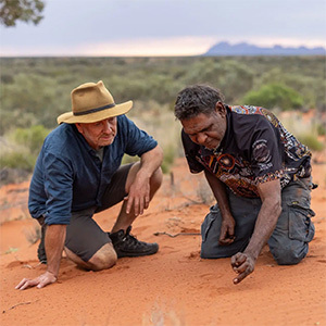 Two men crouch in red sand examining animal tracks